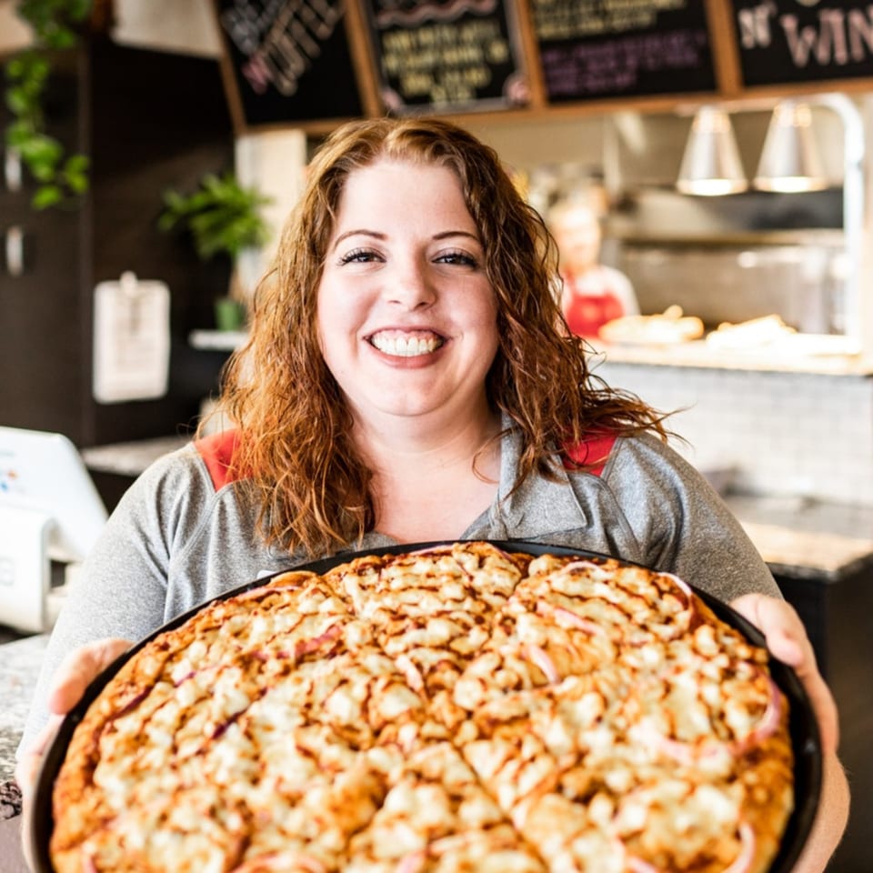 A smiling woman holds a large, freshly baked BBQ chicken pizza with red onions at Slice of Veyo.