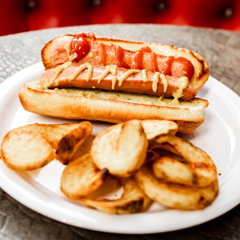 A hot dog with ketchup and mustard, served with a side of potato chips on a white plate.