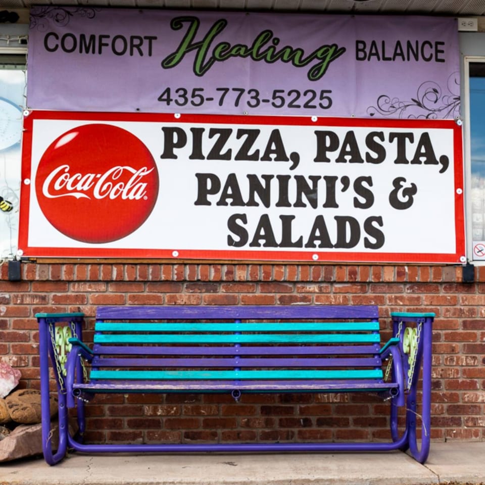 Slice of Veyo's storefront with a sign advertising pizza, pasta, paninis, and salads above a colorful bench.