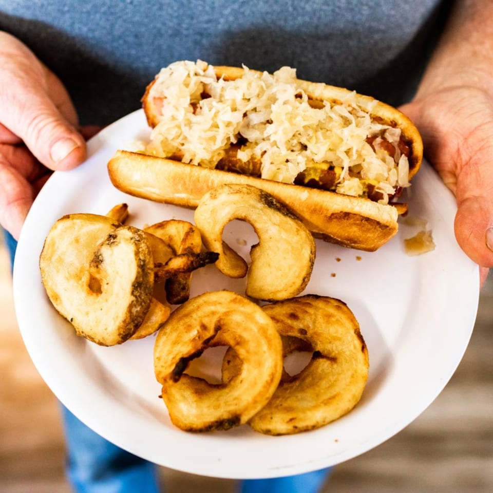 Hands holding a white plate with a hot dog topped with sauerkraut and a side of curly fries.