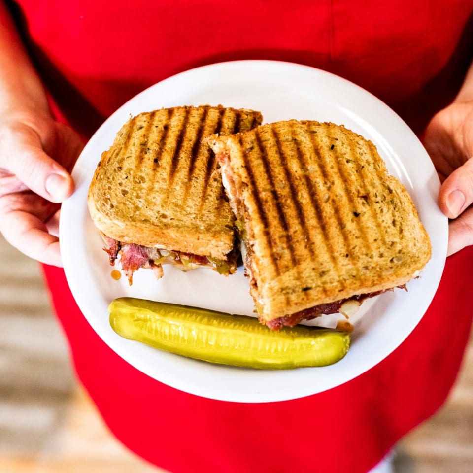 Hands holding a white plate with a grilled sandwich cut in half and a pickle spear.