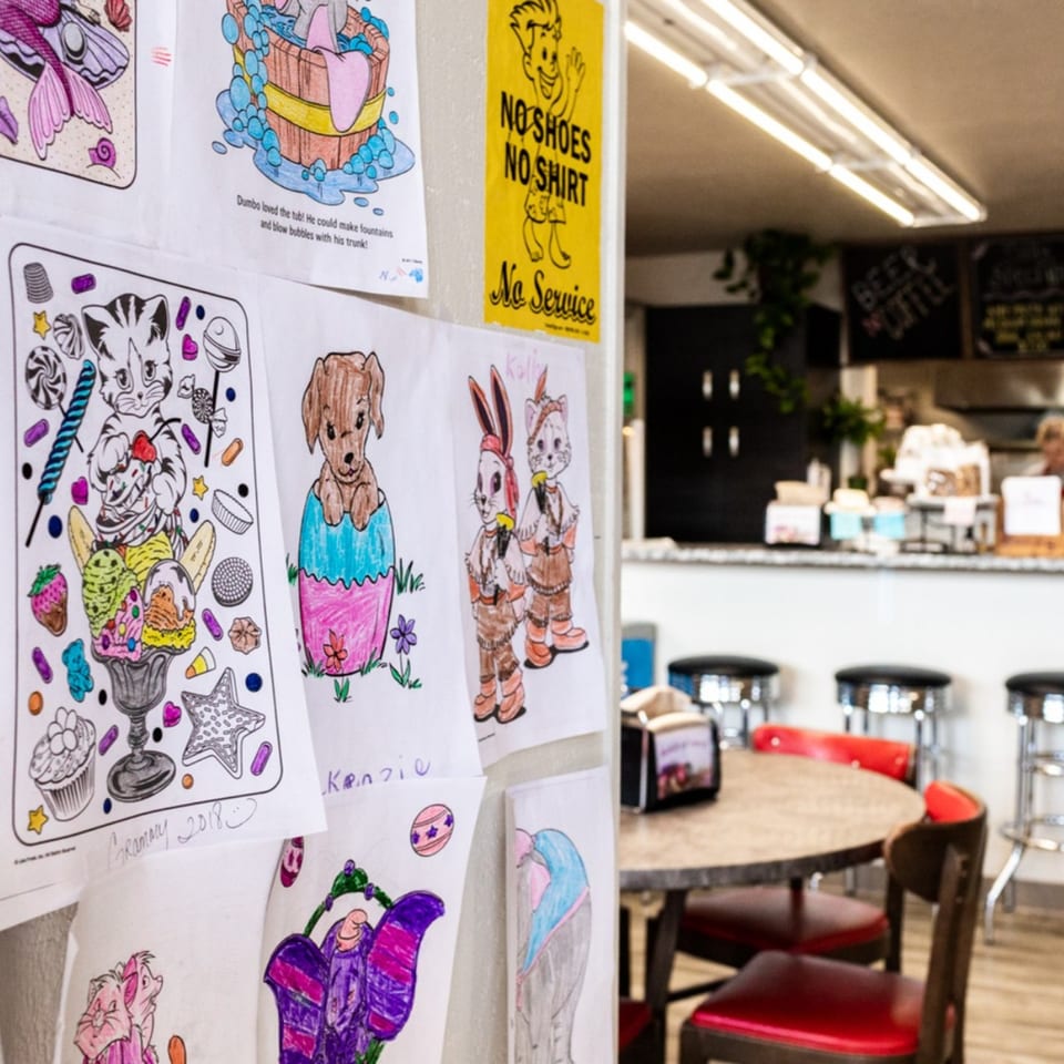 Children's drawings and a "No Shoes No Shirt No Service" sign on a wall, with restaurant seating in the background.