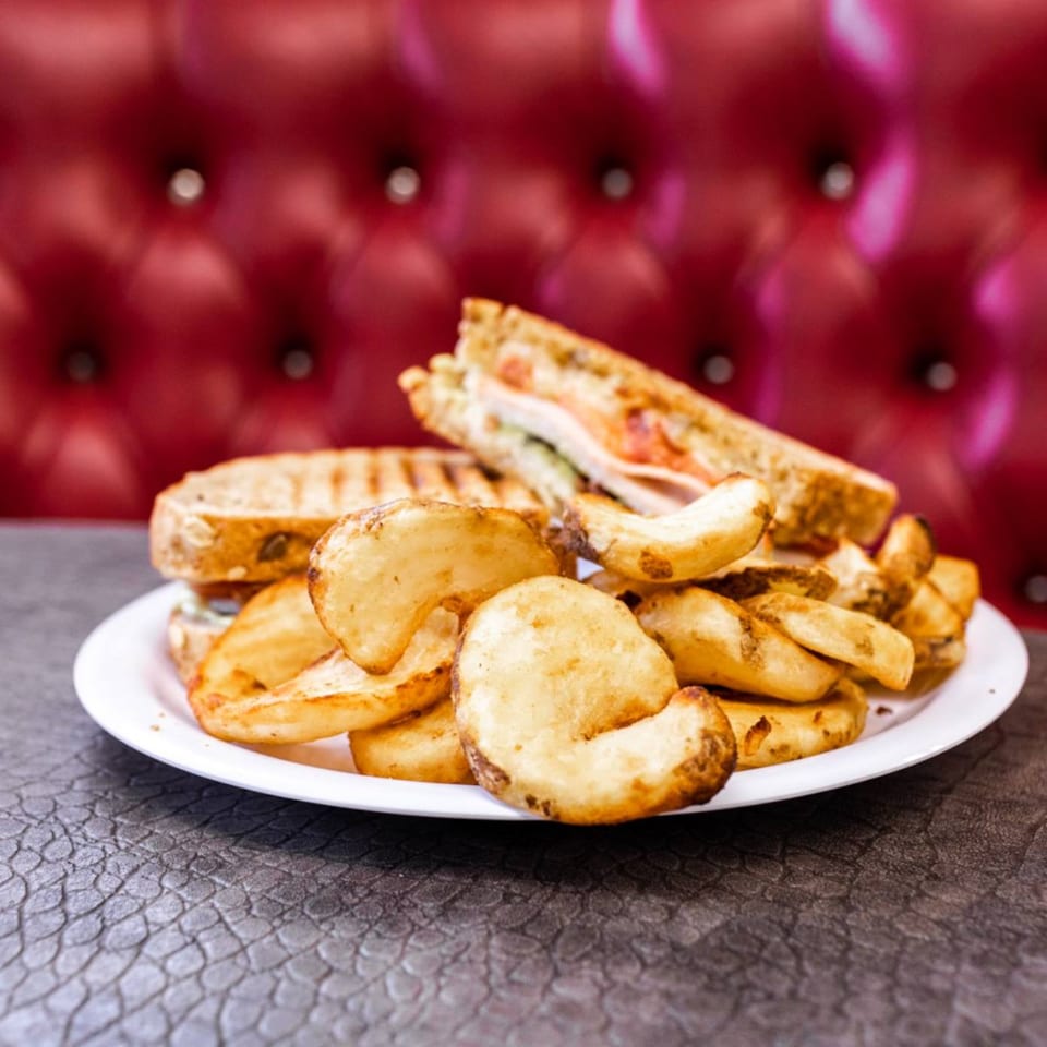 A grilled sandwich and potato wedges on a white plate, with a red tufted booth in the background.
