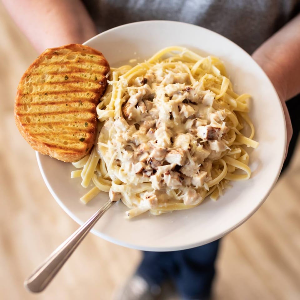 Creamy chicken alfredo pasta with grilled garlic bread on a white plate, held by a person.